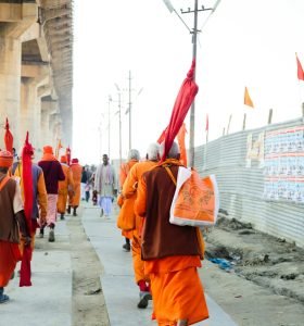 A group of pilgrims in vibrant traditional garb march down an urban street under a bridge.